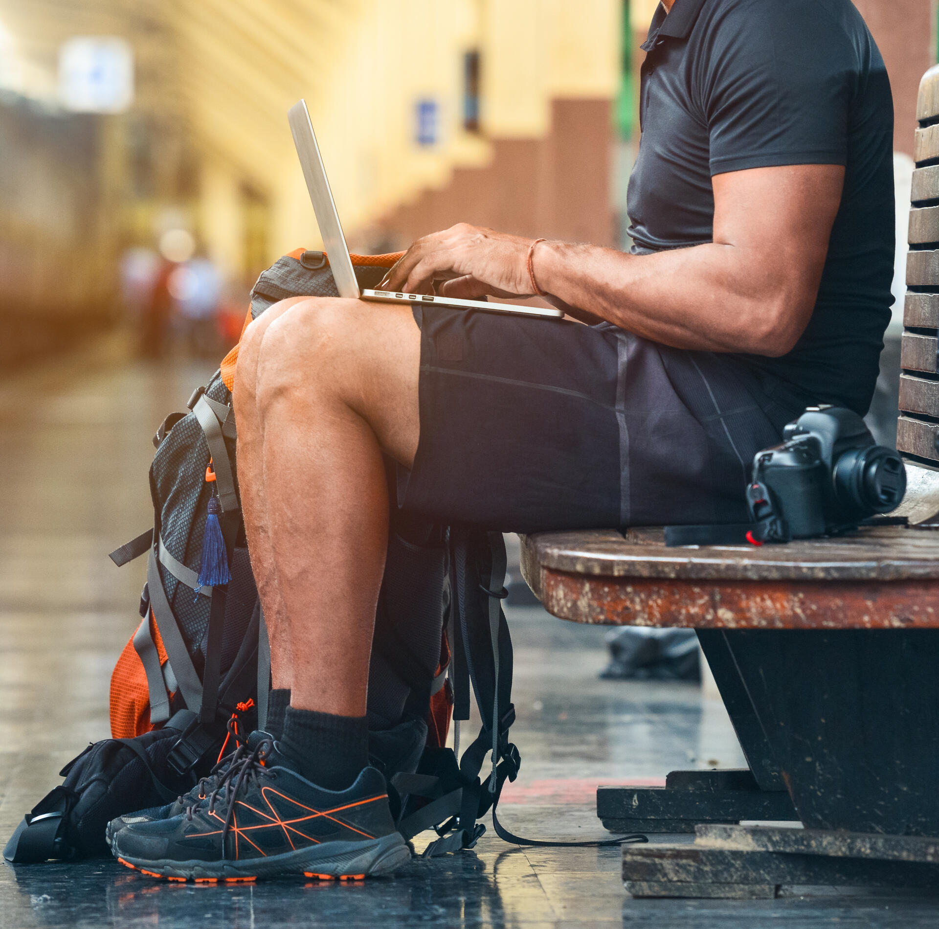 Young man sitting on a bench in a train station, typing on a laptop, camera and backpack beside him.